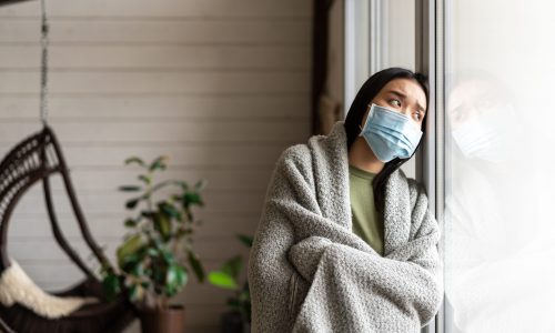 Sick asian woman in medical face mask looking outside window, having flu or covid, staying on quarantine at home.